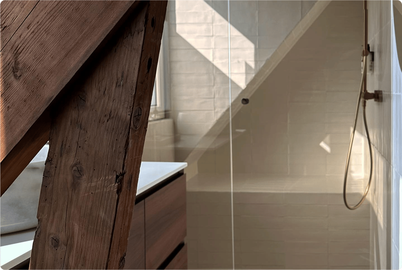 Charaktervolles x²o Bad mit schräger Decke und sichtbarer Holzbalkenstruktur. Rechts eine Dusche mit weißen Fliesen und goldfarbiger Regendusche. Links ist ein Holzbadmöbel unter einem Fenster mit einfallendem Sonnenlicht zu sehen.